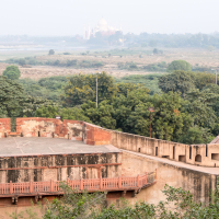 The image depicts an elevated view of historical fortifications with stone walls and a terrace in the foreground, surrounded by green foliage. In the hazy distance, the iconic white dome and minarets of the Taj Mahal in India are visible, blending into the horizon under a light, misty sky.