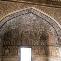Intricately designed historical room with arched ceilings and walls adorned with detailed geometric and floral patterns typical of India. The stone carvings feature symmetrical motifs, and a bright light shines through an open narrow doorway in the center, illuminating the intricate details of the structure.