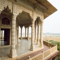 A beautifully ornate pavilion with intricately carved pillars and arches stands elevated, offering a panoramic view of the Indian landscape. The structure, adorned with detailed patterns, overlooks a vast, sunlit expanse of greenery and distant foliage under a clear sky.