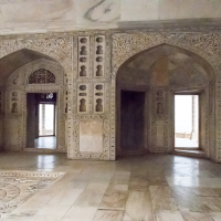 A historic stone room adorned with intricate carvings covers the walls and archways, reminiscent of ancient India. The floor exhibits a detailed, decorative pattern. Light filters in through the doorways and windows, illuminating the delicate craftsmanship of the architecture.