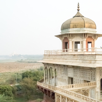 A historical building with an ornate balcony overlooking a sprawling landscape in India. The structure features intricate latticework and a golden dome at its top. The scene extends to a hazy horizon with greenery and fields, highlighting the serene and expansive environment surrounding the edifice.