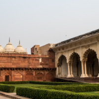 A historical monument in India featuring a large red sandstone wall with three white domes on the left. To the right, an ornate, open pavilion with multiple arched columns is visible. The foreground is occupied by well-manicured green hedges, and the blue sky overhead is clear.