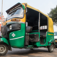 An auto rickshaw with a green and yellow body is parked on the street, partially covering a white car. The auto rickshaw has a CNG label on its front. In the background, the Agra Fort, an iconic building in India made with red bricks and white ornate accents, is visible. A tree and a dog are also present.
