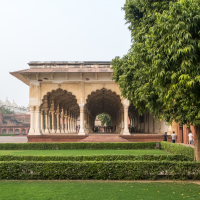 A historic architectural pavilion with arches and columns, reminiscent of India's grand designs, is surrounded by neatly trimmed hedges and green lawns. People walk and sit under the pavilion, shaded by trees. In the background, more domed buildings stand tall beneath a hazy sky.