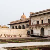 The image shows a historic palace in India with intricate white stone carvings and arches. A domed structure adorned with small decorative spires is visible in the background. A wheelchair ramp leads up to an entrance on the right. In front of the palace, there is a courtyard with patches of greenery.