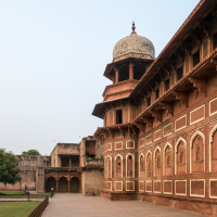 The image shows a section of Agra Fort in India. The structure features ornate red sandstone walls with arched windows and intricate carvings. A large dome is visible atop the building. In the foreground, there is a well-maintained lawn with a single tree to the left under the clear Indian sky.