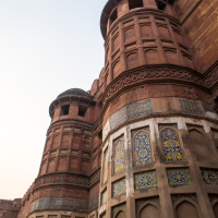 The image shows a close-up of a segment of an ancient fort in India with intricate architecture. The fort, made primarily of sandstone, features high walls and circular towers with decorative arches and patterned ceramic tile work. The clear sky above suggests pleasant weather, inviting travel enthusiasts to explore its rich culture.