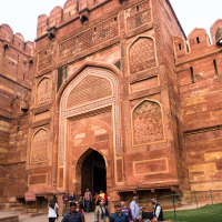 A large, intricate red sandstone gate with detailed carvings and arched entryways forms the backdrop. A group of people, including men and women in various outfits, walk and stand in front of the gate. Set in India, some are taking photos, while others are engaged in conversation or sightseeing.