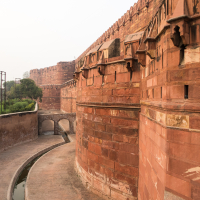 A large, historic red sandstone wall curves towards the viewer, showcasing intricate architectural details reminiscent of India’s rich culture. A curved pathway runs along the base of the wall, leading to an arched bridge. Beyond, lush greenery and trees can be seen under an overcast sky, hinting at possible rain.