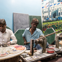 Two artisans work with stone in a workshop. One sits on the left, focusing on detailed hand-painting of circular stone pieces, while the other, in the center, operates a lathe. Surrounded by polished stones and tools, they draw inspiration from a large, vibrant landscape print of India in the backdrop.