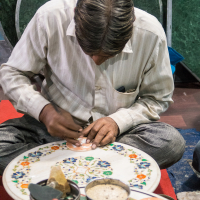 A craftsman is sitting cross-legged on the floor, intricately carving traditional Indian designs into a circular piece of material. He is surrounded by various tools, decorative plates, and small containers. The craftsman is focused on his work, with his head bent down towards the piece.