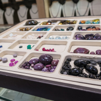 A display case with assorted gemstones on a glass counter. The gemstones, sourced from India, vary in color, including purple, black, red, green, blue, white, and brown and are organized into different sections. Necklaces are hanging in the background out of focus.