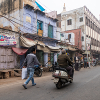 A busy street scene in India with people walking and riding bikes and scooters. The street, quintessentially Indian, is lined with various shops and old buildings, some with peeling paint and advertisements. Electrical wires crisscross overhead. Several vehicles are parked along the roadside.