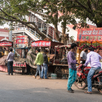 A bustling street scene in India features several food stalls with colorful signs. Vendors and customers interact; two men on a motorcycle are engaged in conversation. A large tree provides shade, and stairs in the background lead up to a building. Various goods are displayed on the stalls, capturing the lively essence of Indian markets.