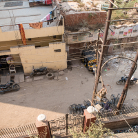 An aerial view of a dirt street in India, bordered by a yellow, multi-storied building with balconies holding clotheslines. The street is lined with parked motorbikes and an auto rickshaw. On the opposite side, a pile of garbage and rubble sits near a brick wall. Power lines crisscross above the scene.