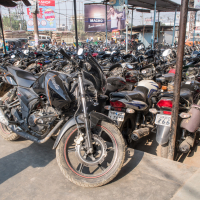A densely packed motorcycle parking lot under a corrugated metal roof, reminiscent of bustling cities in India. Numerous bikes are arranged closely, showcasing a variety of models and colors. In the background, billboards and signs are visible above the lot, with people walking nearby. Sunlight casts shadows across the scene.