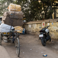 A street scene in India with a cycle rickshaw loaded with large, stacked sacks secured with ropes. A white motorcycle is parked next to a yellow wall adorned with posters and religious images. In the background, a person is seen walking past a row of buildings under a canopy of trees.