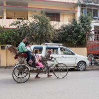 A man pedals a bicycle rickshaw carrying four passengers, including a woman, two children, and a man, along a street in an urban area of India. The background features a white SUV, a red truck, and various buildings with greenery. The rickshaw is in motion with the passengers seated comfortably.