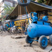 A blue air compressor sits on a dusty street under a makeshift tarp canopy in India. Nearby, bicycles are parked, and several people are seated and standing around. The vibrant backdrop of colorful shops and buildings, with signage in Hindi and a bright yellow and black painted wall, completes the scene.