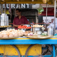 A street vendor in a maroon shirt stands behind his cart selling various food items, including tomatoes, bread rolls, and spices. The yellow and blue cart with large wheels adds vibrant charm to the bustling Indian street. Two customers are partially visible on either side, against a backdrop of restaurant signs and shuttered shops.