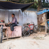 A man is repairing a bicycle on a makeshift workbench in an outdoor, rustic workshop in India. A red bicycle leans against a nearby wall, and a large tire hangs from a wooden structure. A tarp is overhead, providing shade. Surrounding the area are various tools, bicycle parts, and general clutter.