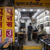 A sewing machine shop with yellow signage and red lettering. Inside, a person stands behind a counter displaying sewing machines, while another sits and one appears to be observing. Various models line the well-lit shelves. An individual, possibly a security guard, is seated outside the shop in India.