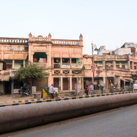 Street scene with old, weathered buildings in pastel colors. People walk and cycle along the sidewalk, and a man in a rickshaw rides by—a typical scene in India. Trees and shop signs are visible, and a large pipe runs along the road. The sky is clear, giving a sense of an early morning or late afternoon setting.
