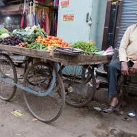 A street vendor sits on a bench beside his wooden cart filled with various fresh vegetables, including tomatoes, greens, and root vegetables. The scene is set in a bustling market area in India, with shops in the background. The ground is littered with debris, and a scooter is parked nearby.