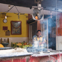 A man stands behind a counter at a street food stall in India, with smoke rising from the cooking stoves in front of him. The stall has a yellow wall with a clock and religious poster. Pots and cooking equipment are on the counter. The background shows kitchen utensils and seating area with green chairs.