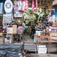 A cluttered shop in India filled with various sewing machines, both new and old, displayed in the entrance and on shelves inside. Two men are seated inside, possibly working or conversing. The shop also has colorful plastic buckets and other household items. A motorcycle is parked in front of the shop.