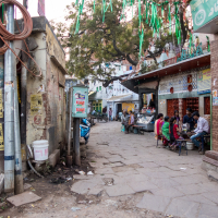 A narrow, bustling street in an Indian marketplace. To the left, an elderly man stands near utility poles and an assortment of wires. The right side features shops, including one with an eye care advertisement in Hindi. In true India fashion, many people are seated, conversing, and engaging in various activities.