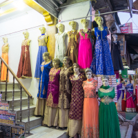The image shows the front of a clothing shop in India, displaying an assortment of colorful traditional dresses on mannequins. Located in a busy market area, the mannequins are arranged on tiered steps, showcasing various styles and vibrant colors, with potential customers and other market stalls visible in the background.
