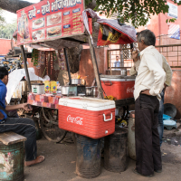 A street food vendor in India operates a small roadside stall. Two men are seen: one seated on a makeshift stool eating and the other standing with his back turned, seemingly in conversation. The stall is adorned with bright signage and various food items, including Coca-Cola coolers prominently displayed.