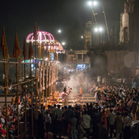 A large crowd gathers at a riverbank in India during a nighttime ceremony in Varanasi. Priests perform rituals under brightly lit, ornate canopies with flaming torches, as smoke fills the air. The audience is seated and standing, watching intently. Tall flags and historical structures are visible.