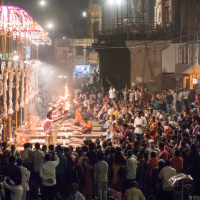 A large crowd gathers to watch a traditional Hindu ceremony along the riverbank in Varanasi, India. Priests in white and saffron robes hold burning lamps while performing the evening Ganga Aarti. The scene is illuminated by bright lights and decorated structures, showcasing the cultural richness of India.