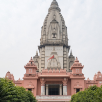 Image of Kashi Vishwanath Temple, India, featuring a tall, ornate spire with intricate architectural details. The temple's entrance shows pink walls, multiple smaller spires, and an open passage. The pathway leading to the entrance is lined with trees on both sides, under a cloudy sky.