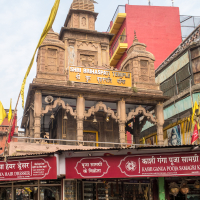 A bustling street in India with a historic temple, the Shri Brihaspati Temple, prominently displayed above shops, including a hairdresser and a puja samagri store. The temple features intricate carvings and multiple spires. Flags and signs in Hindi adorn the buildings, with people walking nearby.