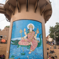 A large mural depicting a Hindu deity riding a crocodile adorns a cylindrical water tower in India. The deity holds a trident, conch shell, and lotus flower. The scene includes water with fish and turtles. Around the base, people are gathered, and steps leading to the river are visible in the background.