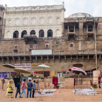 A group of people stand and walk on wide stone steps in front of Manmandir Ghat in India. Above them, ornate architecture blends white and brown stone with intricate carvings. Vendors have umbrellas set up, displaying goods and textiles. A sign in Hindi and English reads "Manmandir Ghat.