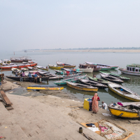 A riverside view in India showcases numerous colorful wooden boats docked along the shore of the Ganges River. Two people stand near a small yellow boat on a sandy bank. The water stretches to the horizon under a hazy sky, with steps in the foreground leading down to this serene scene.