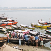 A group of men in casual clothing, some shirtless, use ropes to pull a boat ashore at a busy riverside docking area in India. Numerous colorful wooden boats are tied up nearby on the calm river, which fades into a misty horizon with distant trees visible. The scene is bustling and industrious.