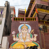 A large mural of the Hindu deity Ganesha is painted on a building wall in India. The artwork features Ganesha seated, with multiple arms holding various objects. Above the mural, the word "GANPATI" is written. The building is decorated with colorful flags and sits under a partly cloudy sky.
