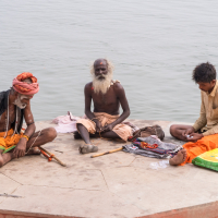 Three men sit cross-legged on a hexagonal concrete platform by a body of water in India. The man on the left has an orange headwrap and wears beaded necklaces. The central man, older with a white beard, sits with a piece of cloth on his lap. The third man, in beige and orange, is surrounded by various items.