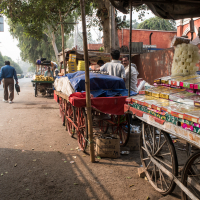 A street market scene in India shows various vendor carts lined up along a tree-shaded sidewalk. The carts are covered with assorted goods, including sweets and snacks. People walk and shop along the street. In the background, a green road sign indicates directions to a fort and Taj Mahal.