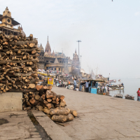 A riverside ghat in India is bustling with activity, featuring piles of logs in the foreground, likely for use in rituals. People gather on the steps and along the water's edge where boats are anchored. Historic buildings and temples with intricate architecture stand in the background under a hazy sky.