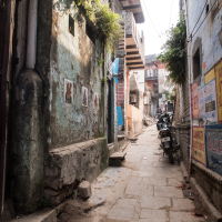 Narrow alleyway between aged, weathered buildings with peeling paint and wall posters, reminiscent of old India. Several motorbikes are parked on the right side, near colorful signs and advertisements. Stone-paved ground with steps leading to doorways. Small plants grow on ledges above. Distant light suggests broader street.