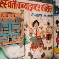 A mural on a wall in India depicts two schoolgirls in uniform and Mickey Mouse. The girls, one holding books, wear red skirts and white shirts, and carry schoolbags. The mural includes faded text in Hindi and English, and shows a building sketch labeled "SKIC." The setting appears to be a school entrance.