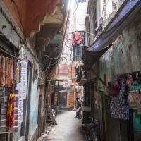 Narrow alleyway in a densely built, urban area in India. Shops lined up on both sides sell colorful trinkets and textiles. Overhead, wires crisscross between buildings. The alley is paved with stone slabs. Sunlight filters in, casting shadows. A bicycle is parked along the right side ahead.