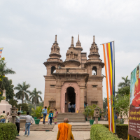 A Buddhist monk dressed in vibrant orange robes walks towards a temple in India, with multiple small towers and a large central spire, holding a walking stick. The ornately adorned temple is surrounded by greenery and people. Two tall, colorful flags flank the pathway leading to the entrance.