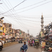 A busy street scene in an urban area of India with numerous shops lining both sides. People walk, cycle, and ride auto-rickshaws amidst bustling activity. Overhead, power lines crisscross the sky. On the right, a tall minaret stands out prominently among the buildings. Vehicles and motorcycles are parked along the sides.